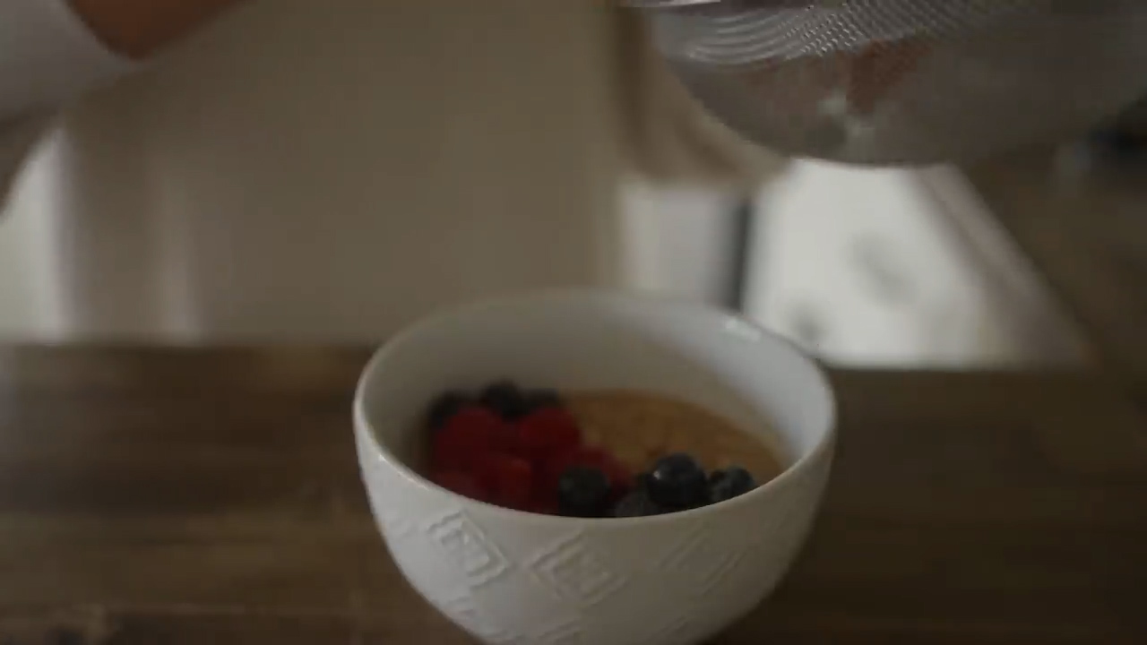 a white bowl containing oatmeal, blueberries and raspberries sits on a wooden surface. a woman's hands can be seen holding a sieve above the bowl, as though she is about to sprinkle something like sugar over the contents. the focus is on the food, with the woman and the background slightly out of focus.