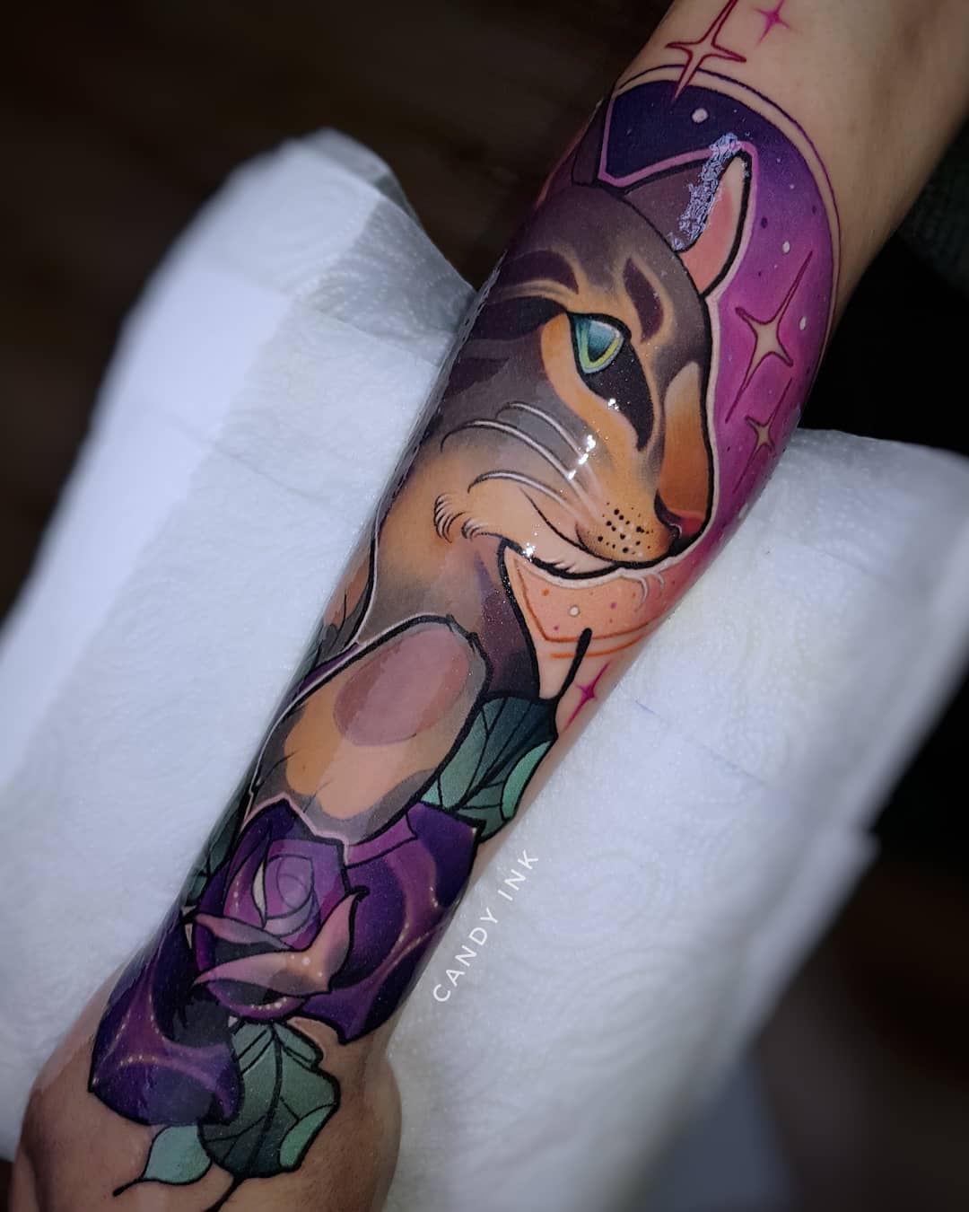 a person's forearm with a fresh neo traditional tattoo of a cat. the cat is gray with orange stripes and green eyes, set against a backdrop of a purple galaxy with stars. below the cat is a large purple rose. the tattoo is detailed and colorful.