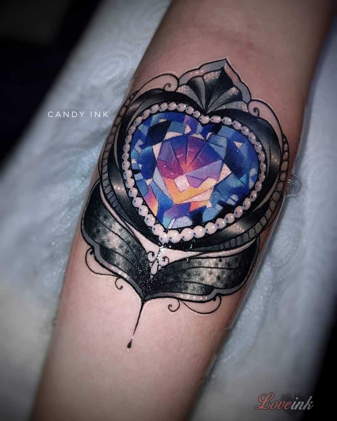 a tattoo of an ornate heartshaped jewel on someone's inner forearm.  the top of the heart jewel is a sapphire blue color that fades to a pink and orange at the bottom. the heart is surrounded by white pearls and an elaborate black and gray design.