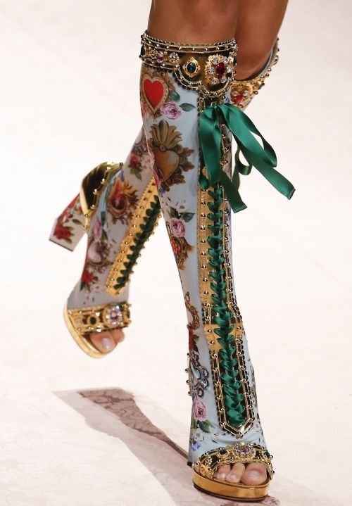 closeup shot of a model's leg wearing a pair of striking dolce & gabbana boots. the boots are highheeled and feature a light blue floral print overlaid with intricate gold baroque embellishments. a green satin ribbon laces up the sides, adding a contrasting element to the design. the platform heel and opentoe style are distinctly visible. the overall aesthetic is luxurious and opulent, suggestive of a highfashion runway presentation.