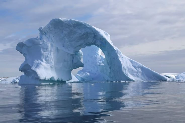 an iceberg floats in the ocean with its reflection visible in the water. the sky is cloudy and the iceberg has a large arch carved into it.