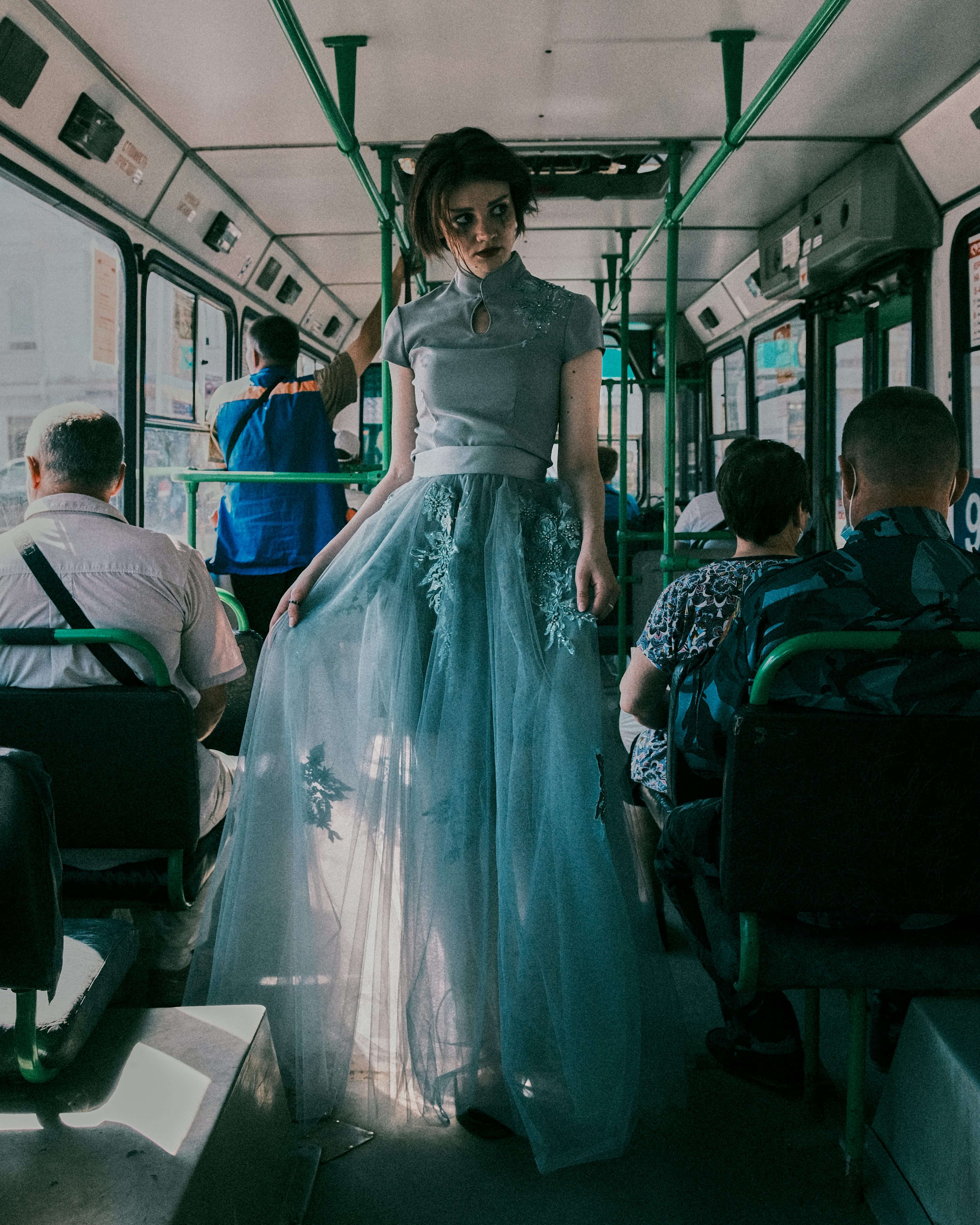 a woman in a flowing light blue tulle dress stands in the aisle of a crowded public bus. the dress is long and elegant, creating a stark contrast to the utilitarian setting.  the overall mood is moody and somewhat mysterious, enhanced by the dimly lit interior of the bus and the woman's somewhat pensive expression. the other passengers are blurred, drawing the focus to the woman in the dress. the image has a vintage, filmlike quality.