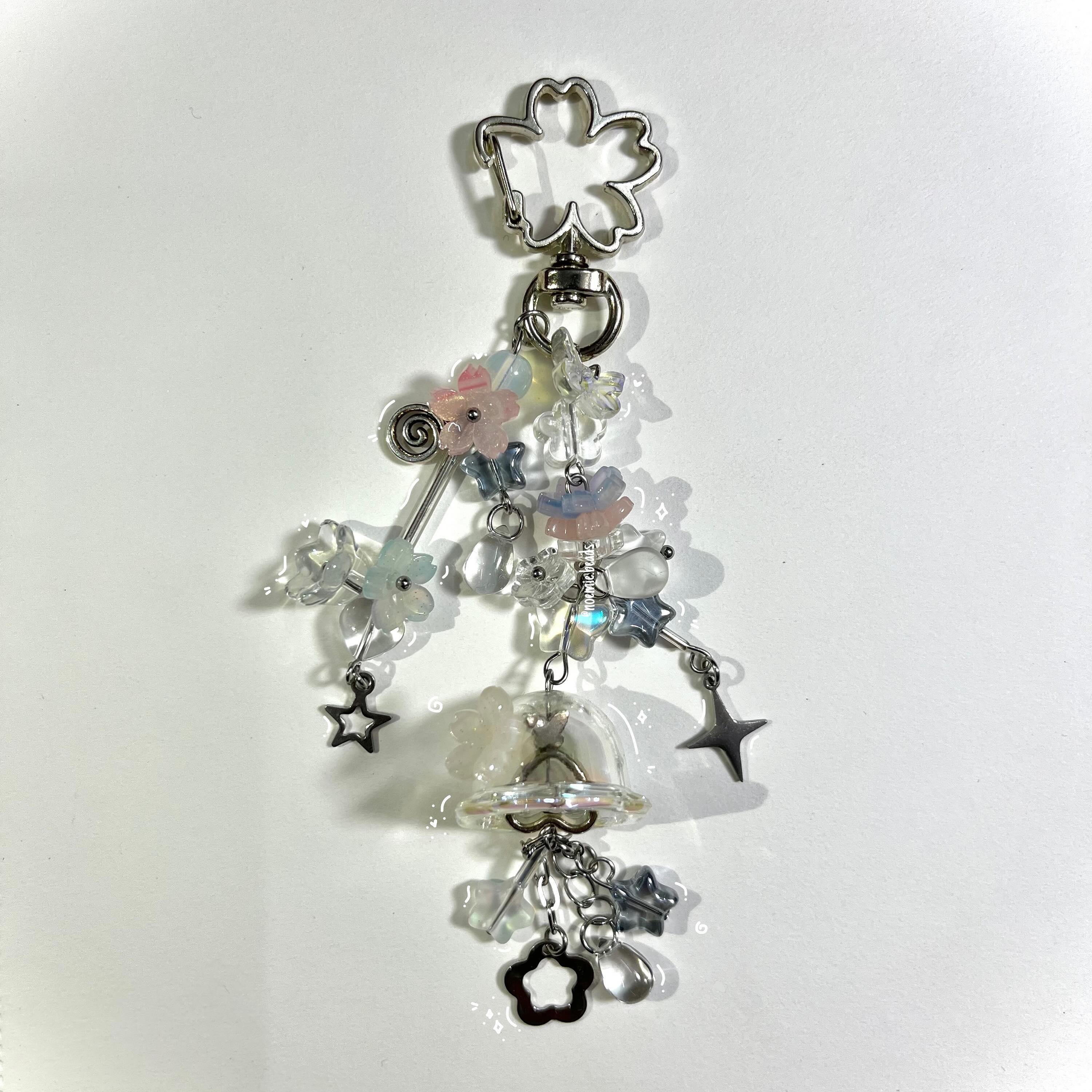 a closeup studio shot of a handmade keychain featuring an array of transparent, iridescent, and pastel beads and charms, including flowers and stars, all hanging from a silver flowershaped clasp.
