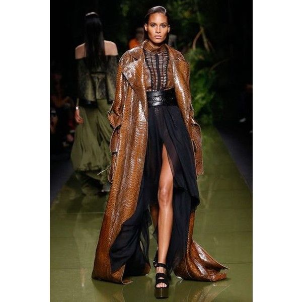 a female model walks down the runway in a stylish outfit. she's wearing a long, brown snake skin coat over a long black dress with a slit. she's wearing a black belt and black high heels. the model is posing for the camera, with a serious expression on her face. the background is blurry and out of focus, but you can see other models and audience members in the background.