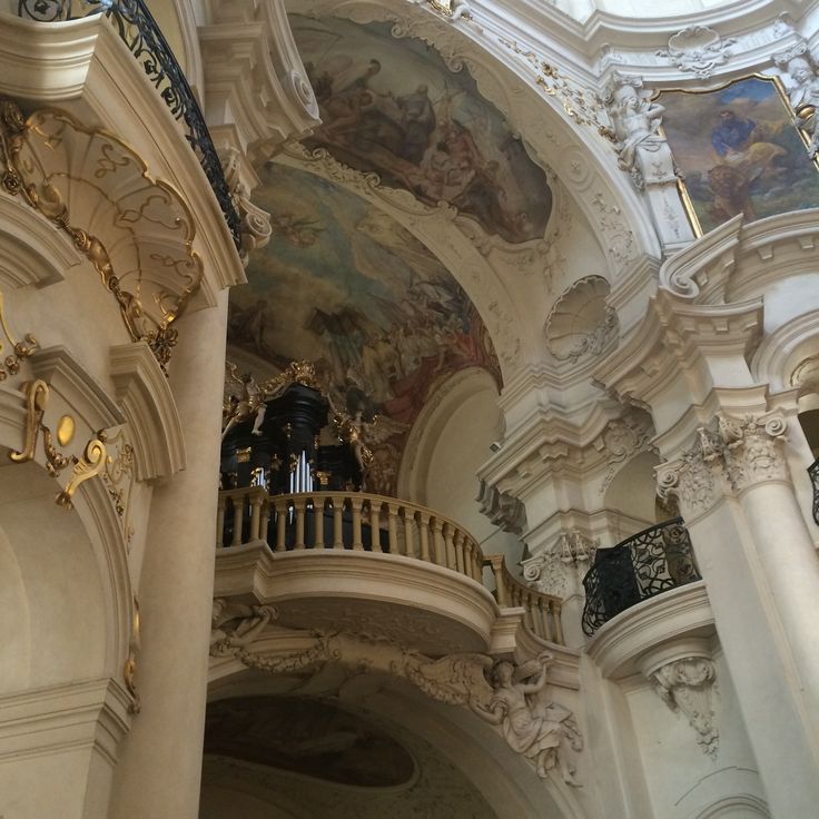 the image shows a stunning interior view of a baroquestyle church.  the focal point is an elaborate organ loft situated within a complex network of arched recesses, columns, and ornate stucco work.  the ceiling is adorned with a large fresco depicting religious scenes, while the walls are richly detailed with gold leaf accents and sculptural angel figures.  the overall aesthetic is one of grandeur, elegance, and rich historical significance, typical of european baroque architecture.  the color palette is predominantly soft creams and beiges.