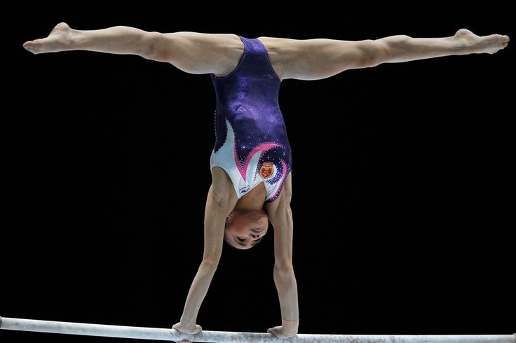 a female gymnast performs a handstand on the parallel bars. the gymnast is wearing a purple, pink, and white leotard. the background is black.