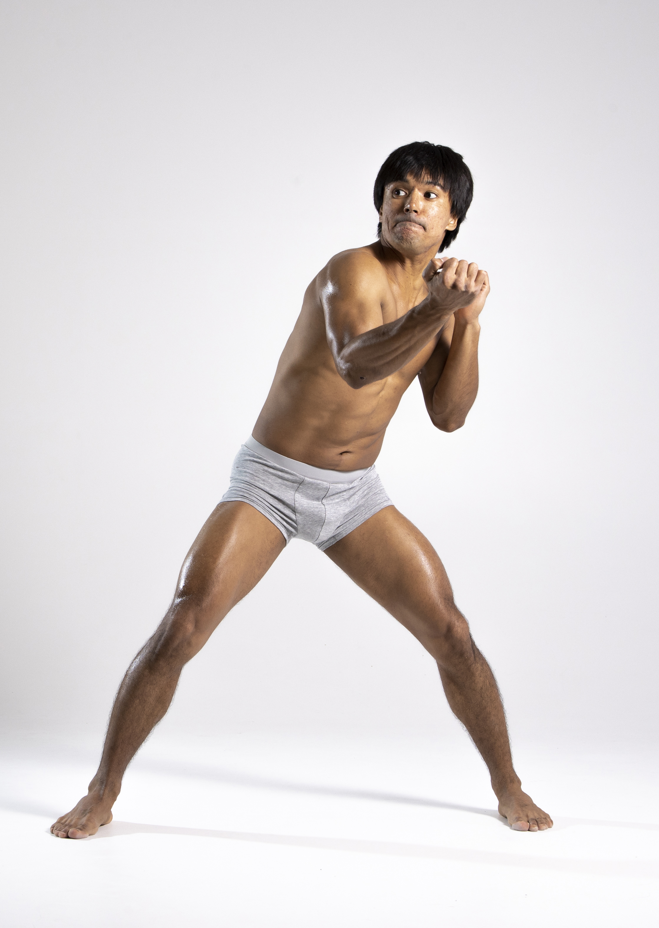 a studio shot of a muscular man in a fighting stance. he is wearing gray boxer shorts and is barefoot. his skin appears oiled and he has dark hair. he is looking off to the side with a tense expression.