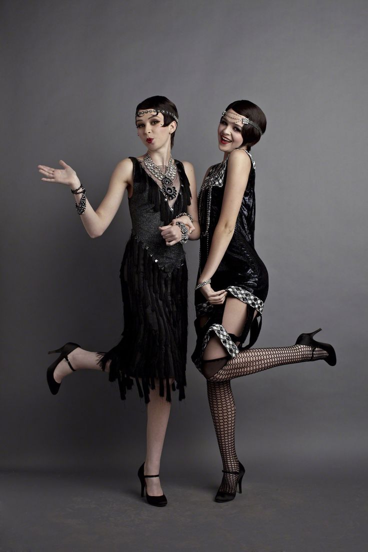 two young women in flapper dresses from the 1920s pose together in a studio setting. the woman on the left is looking towards the camera and is smiling. the woman on the right is looking at the woman on the left. both women are wearing black flapper dresses, black high heels, and jewelry. they are also both wearing a headband with beads, and stockings. the woman on the right has fishnet stockings.