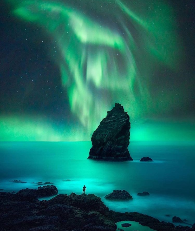 a scenic view of the northern lights over the ocean, with a rocky landscape and a person standing on the rocks. a large rock formation stands in the water with the aurora behind it.