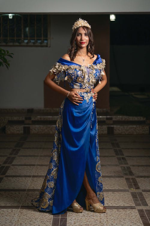 a young woman in a stunning blue gown stands elegantly in a building. she's wearing a gold crown and heels, and her pose exudes confidence and sophistication. the dress is intricately designed, featuring gold embellishments, and its flowing skirt contrasts with the fitted bodice. the setting appears to be an indoor space with tiled flooring, suggesting a formal event or photoshoot. the lighting is dramatic, casting shadows and highlighting the details of the dress and the woman's features.