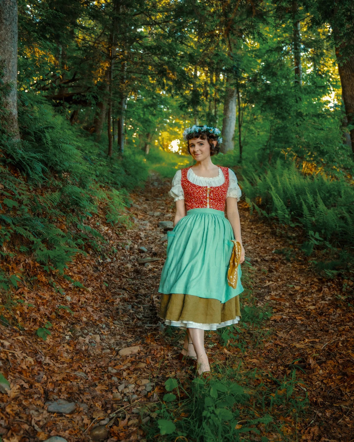 a young woman, adorned in a traditional dirndl dress and a flower crown, strolls along a sundappled forest path. the path is covered in fallen leaves, suggesting autumn or fall.  the surrounding forest is lush and green, with tall trees creating a canopy overhead.  the woman's dress is primarily teal/aqua and brown, with a red bodice. she carries a small folded cloth in her hand. the overall scene evokes a sense of fairytale or whimsical charm.