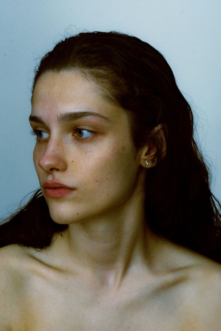 the image is a closeup portrait of a young woman with long, dark brown wavy hair. her skin is pale with visible freckles. she is wearing minimal makeup and has a calm, slightly pensive expression. the woman is shown from the shoulders up, with her bare shoulders visible.  the background is a plain, lightgrey, creating a minimal and elegant effect that focuses attention on her features.  the overall mood is serene and natural.