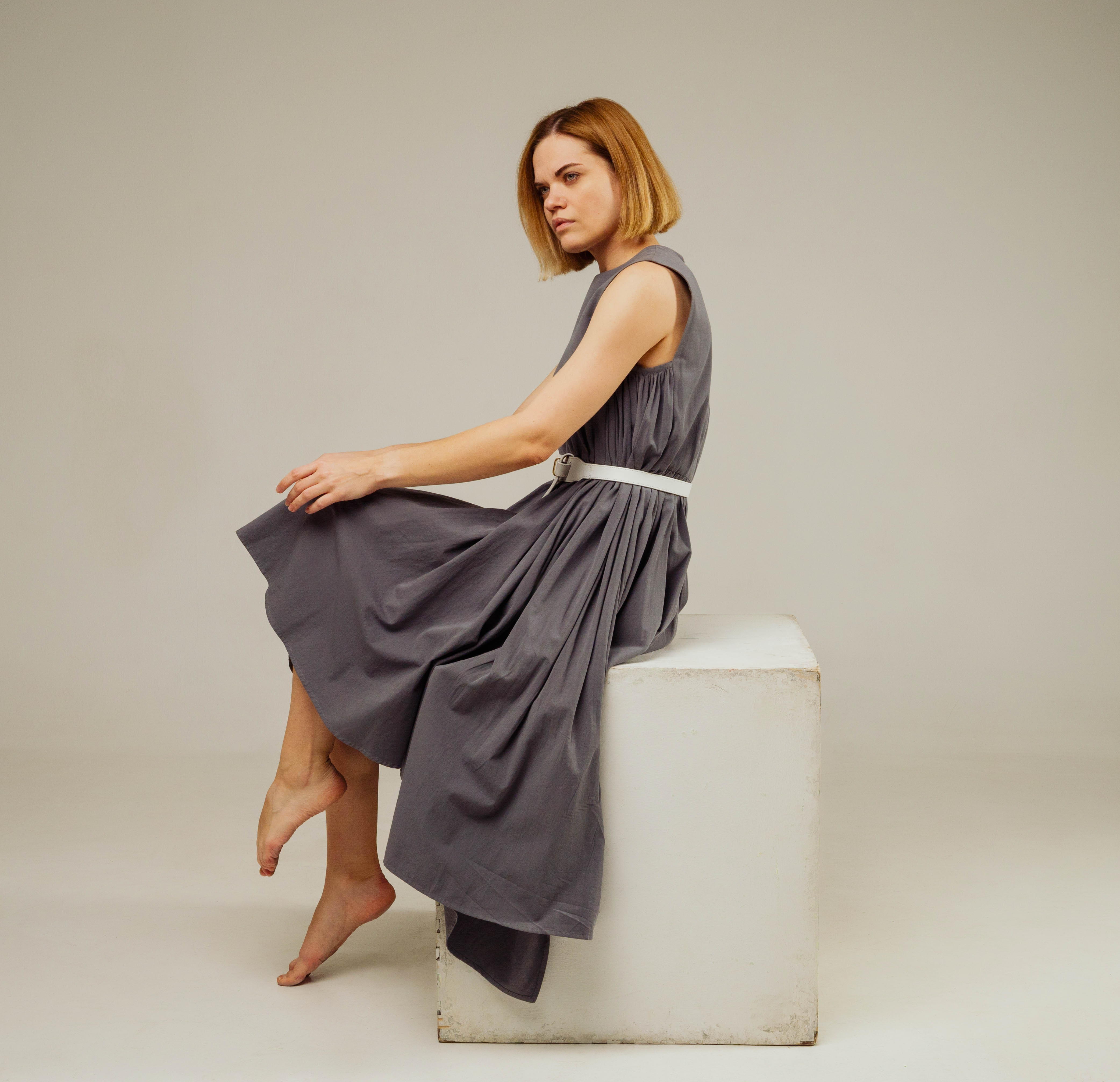 a woman wearing a gray dress with a white belt is sitting on a white cube in a studio. she is looking to the side and has a neutral expression. the lighting is soft and the background is a light gray.