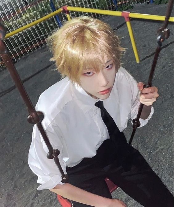 a young man with blonde hair and red eyeshadow wearing a white button down shirt and black tie sits on a swing in a playground at night. he looks at the viewer, lit by artificial lighting.