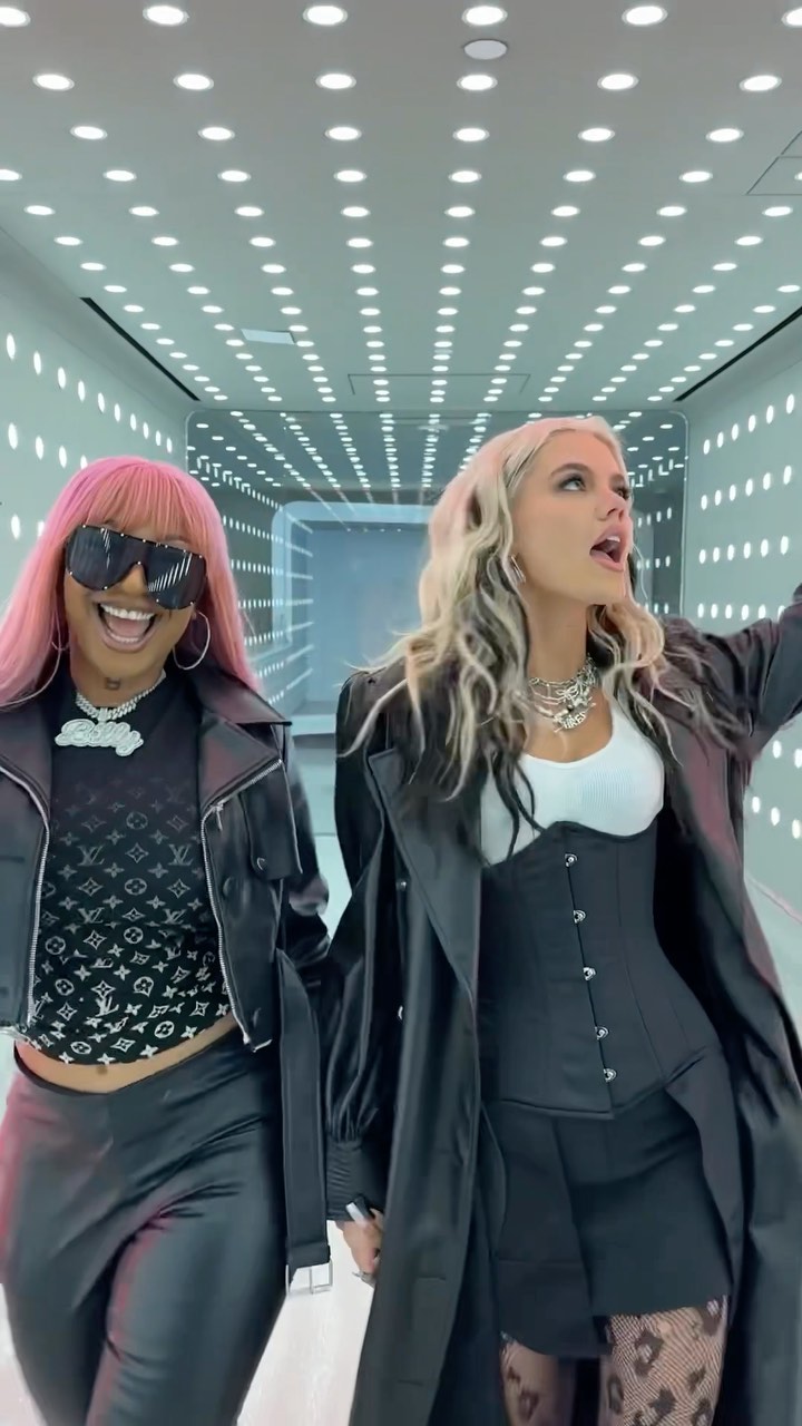 two women are holding hands in a futuristiclooking hallway with rows of lights on the ceiling and walls. the woman on the left has pink hair, large sunglasses, a silver necklace, a black leather jacket over a cropped top with a pattern, and black pants. the woman on the right has blonde hair with dark streaks, multiple silver necklaces, a black coat over a black corset and white top, a black miniskirt, and patterned tights.