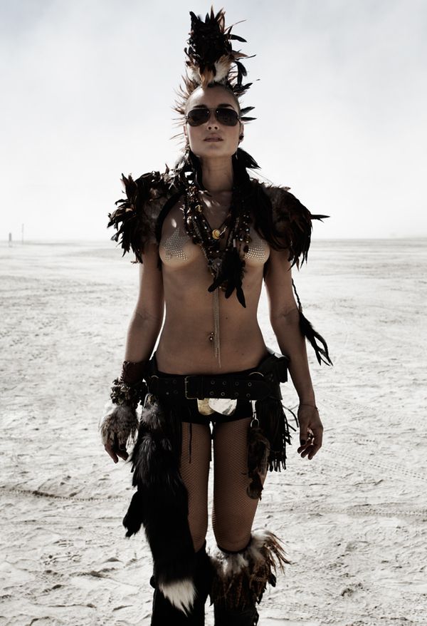 a woman wearing a black and brown feathered outfit poses in the desert. she is wearing a headband with feathers and sunglasses. she is looking at the camera with a serious expression.