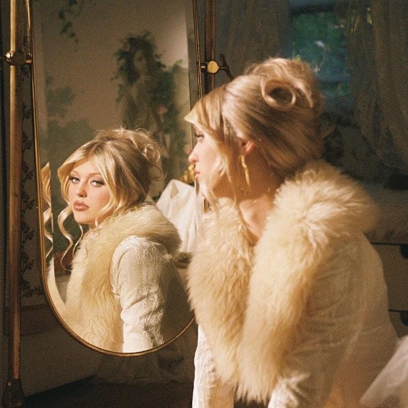 a woman with blonde hair styled in an updo is looking at her reflection in an antique mirror. she is wearing a white dress with a large fur collar. the image has a vintage aesthetic.