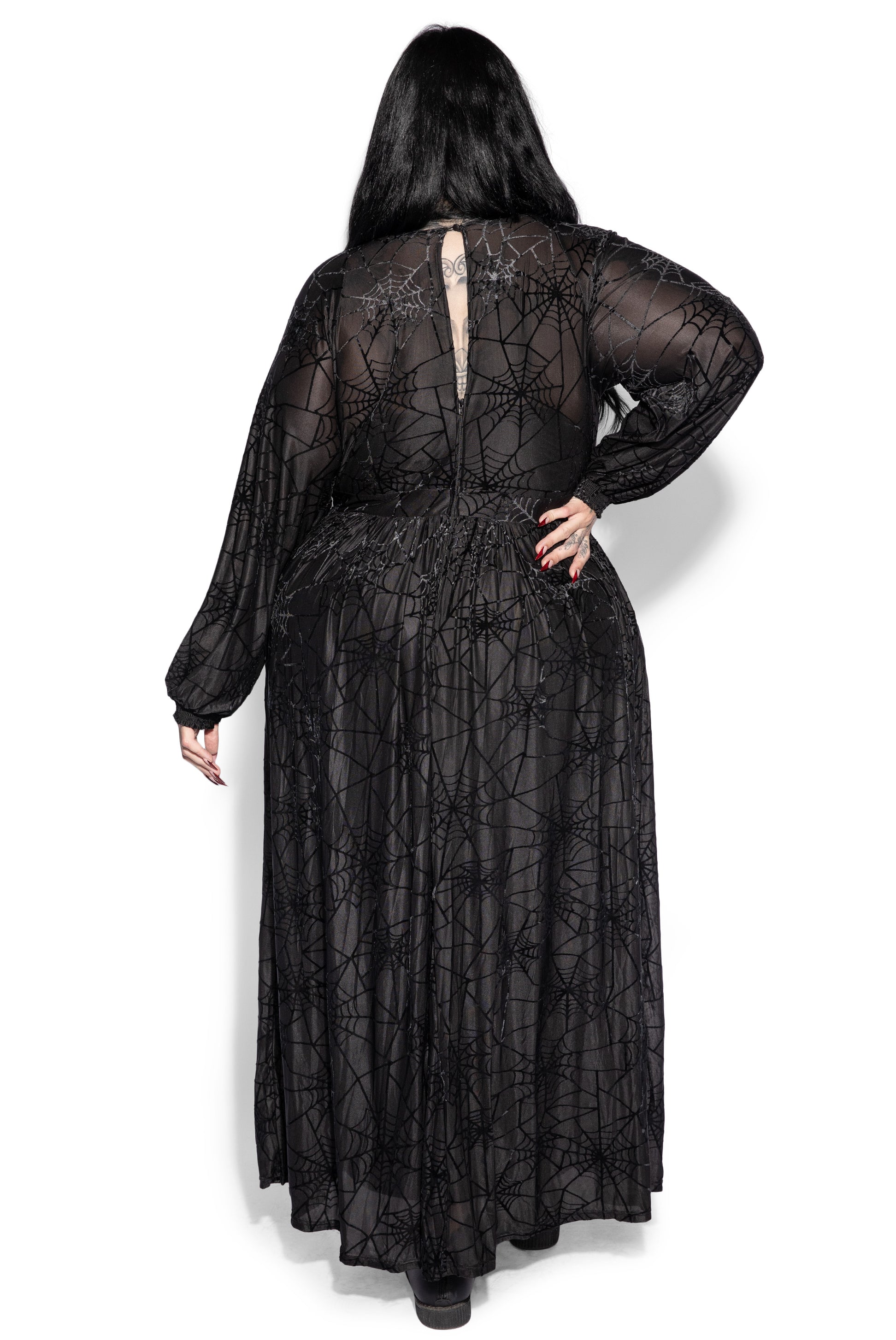 a rear view of a plussize model wearing a long black maxi dress with a spiderweb pattern. the dress is made of velvet and has long sleeves. the model is standing on a white background.