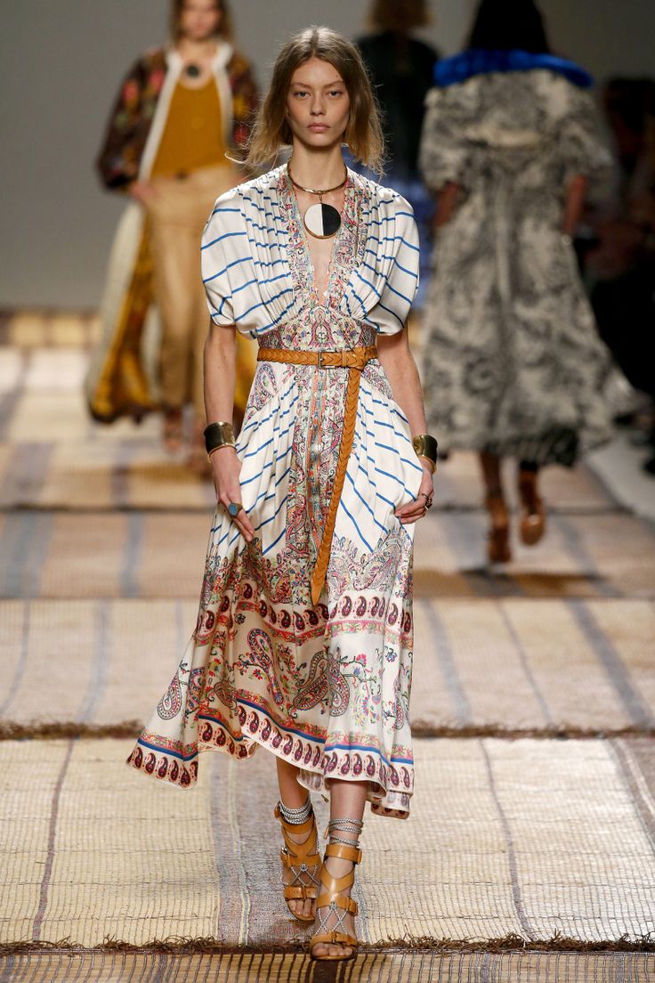 a fashion model walks the runway wearing a beautiful bohemianstyle maxi dress. the dress is white with blue stripes and a paisley print.  it has short puffed sleeves and is cinched at the waist with a brown leather belt. she is accessorized with a large circular necklace, several bracelets, and brown leather sandals. her hair is styled in a bob. two other models are visible in the background, blurred, suggesting a fashion show setting. the runway floor has a textured matting. the overall aesthetic is summery, casual, and chic.