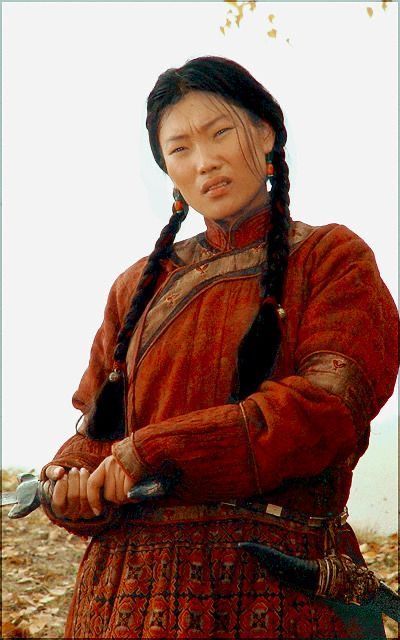 this image is a portrait of a woman with long black braids dressed in traditional warrior clothing. she is standing in a field of autumn leaves and holding a knife with a serious expression on her face. she is dressed in a red and orange outfit with a brown leather belt and a knife strapped to her hip.