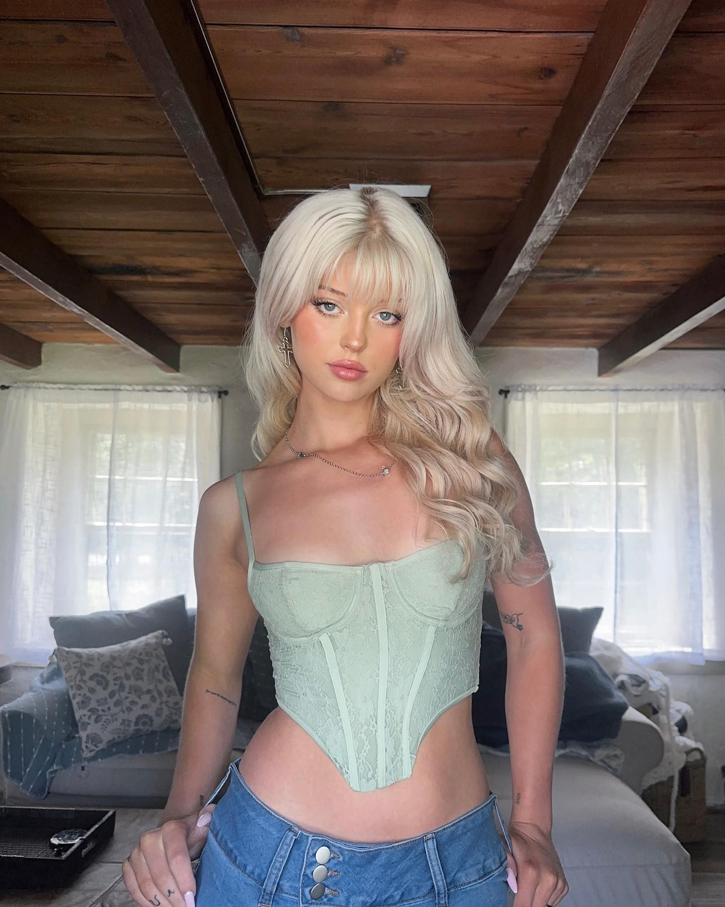 a young woman with blonde hair and bangs is posing in a light green corset top and low rise jeans. she is indoors, likely in a house with a wooden ceiling and large windows. the background includes a sofa with pillows. she is wearing jewelry including a necklace and earrings and has tattoos on her finger and arm. she is looking directly at the camera.