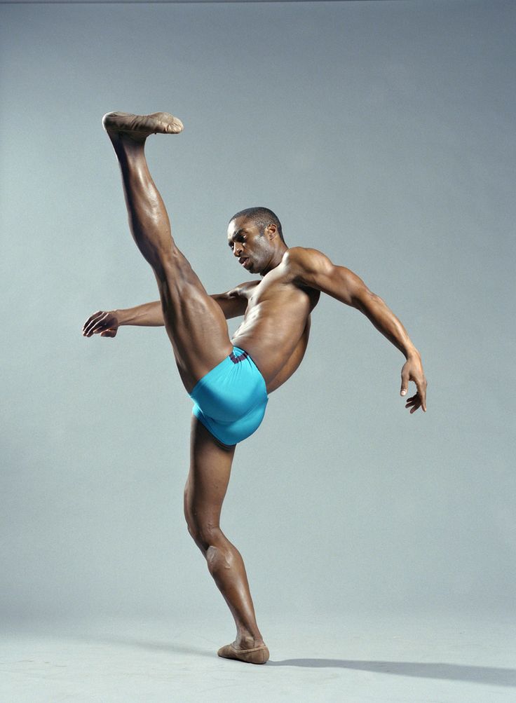 a black male dancer with a muscular physique is posing in a blue dance brief. he is stretching his leg up in a ballet pose. the background is grey and there are no other props or people in the photo. the dancer is looking down at his leg,  he appears to be concentrating on his pose and  he looks fit and strong. the photo is taken in a studio, it is a clean and sharp image.