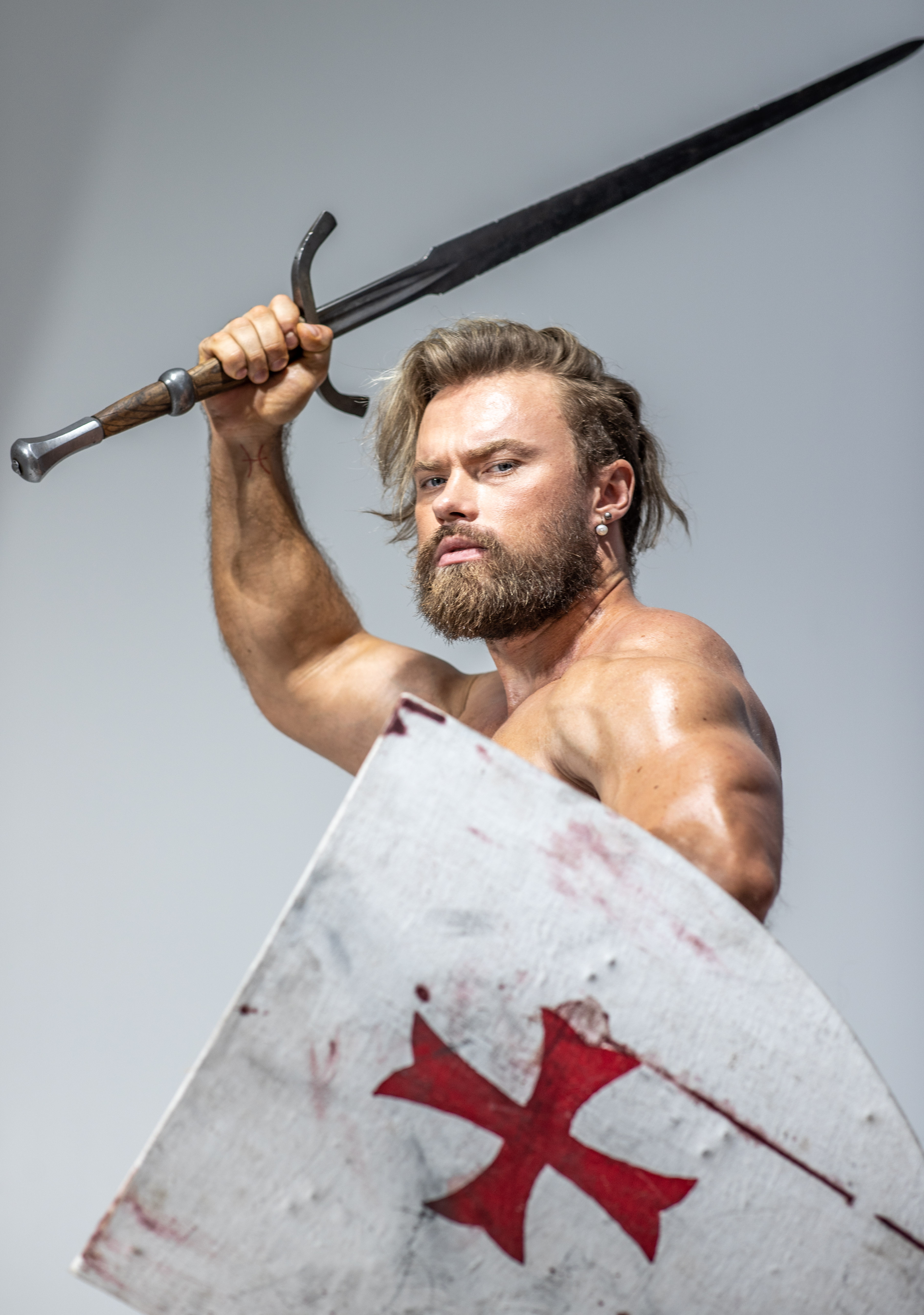 a muscular man with long blond hair and beard, poses barechested against a plain white background. he brandishes a long sword above his head with his right hand while holding a shield emblazoned with a red cross in front of him with his left. he appears to be a warrior from medieval times, possibly a knight.