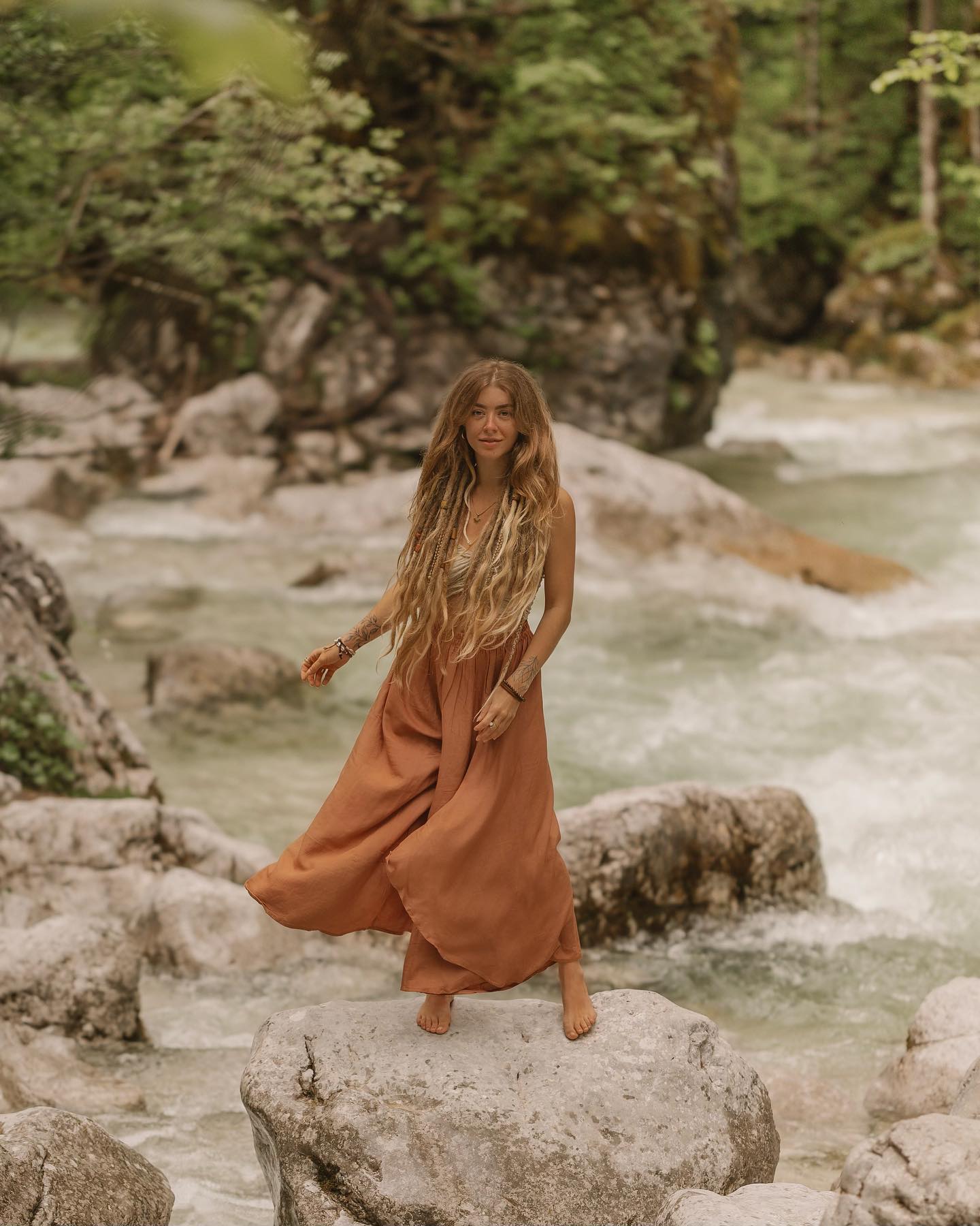 the image depicts a young woman with long blonde dreadlocks wearing a flowing terracottacolored wideleg dress or pants. she is standing barefoot on a large, lightgrey rock in a shallow mountain river. the background features a lush green forest and the rushing water of the river. the overall mood is serene and peaceful, suggesting a sense of freedom and connection with nature. the style is bohemian or hippieinspired, reflecting a relaxed and natural aesthetic. the lighting and color palette create a warm and inviting atmosphere.