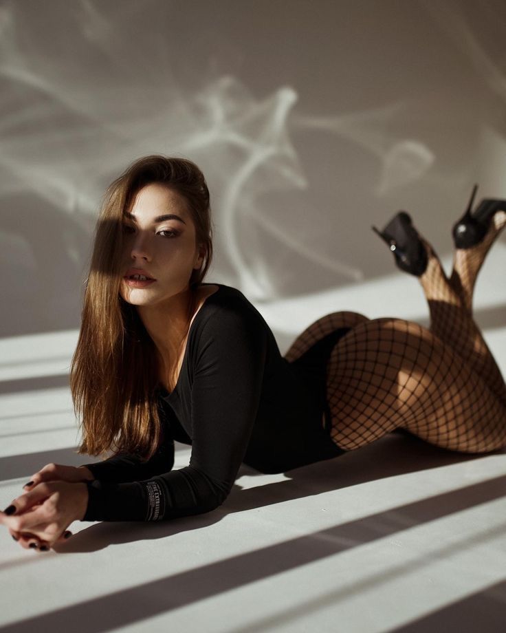 a young, attractive brunette woman with long hair is lying on a white floor, wearing a black bodysuit and fishnet stockings. she is wearing high heels and her pose is sensual and alluring. the image is a studio shot with dramatic lighting and shadows that emphasize her curves and features. the overall aesthetic is fashionforward and glamorous.