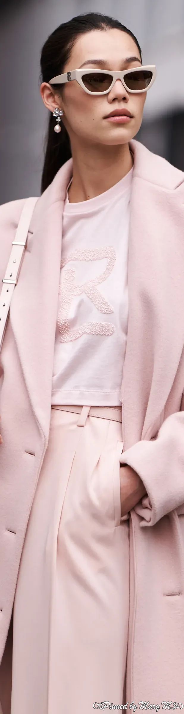 a stylish woman is captured in a fulllength outdoor shot. she's dressed in a chic, monochromatic pink outfit: a long, doublebreasted coat, matching highwaisted trousers, and a simple pink top adorned with an embroidered logo. the ensemble is elevated with pasteltoned cateye sunglasses, delicate earrings, and a cleanlined white handbag. the overall style is minimalistic yet sophisticated, suggesting elegance and a refined fashion sense.  the setting is subtly visible and appears to be an urban environment.