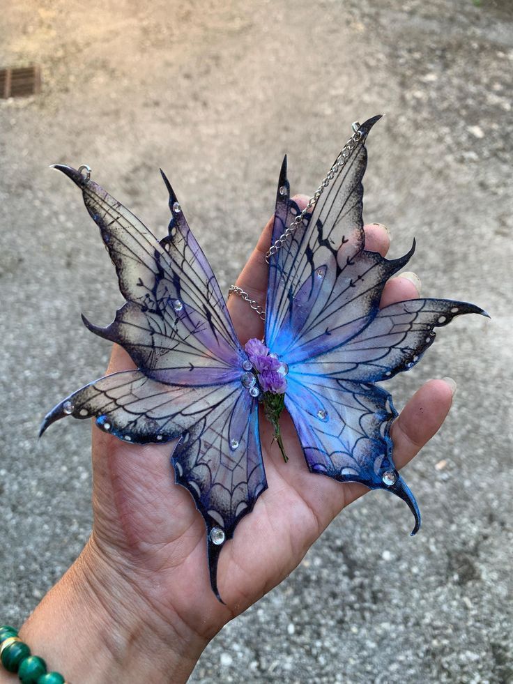 a closeup shot of a person's hand holding a pair of iridescent fairy wings or butterfly wings. the wings are primarily purple and blue, with black accents and rhinestones. a chain connects the two wings at the center, where a small purple flower is also attached. the background is a blurry outdoor scene.