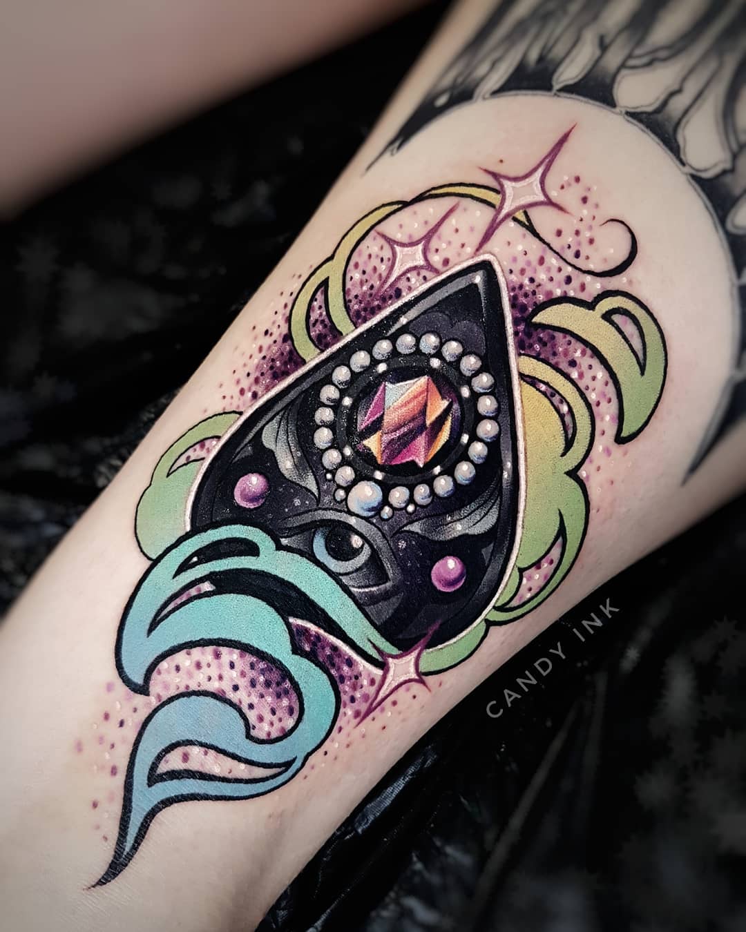 a neotraditional illustrative tattoo of a planchette on someone's forearm. the planchette is black with a large iridescent crystal or gem in the center, surrounded by white dots. it's surrounded by a green, teal, and yellow design of smoke and sparkles. this tattoo was done by candy ink.