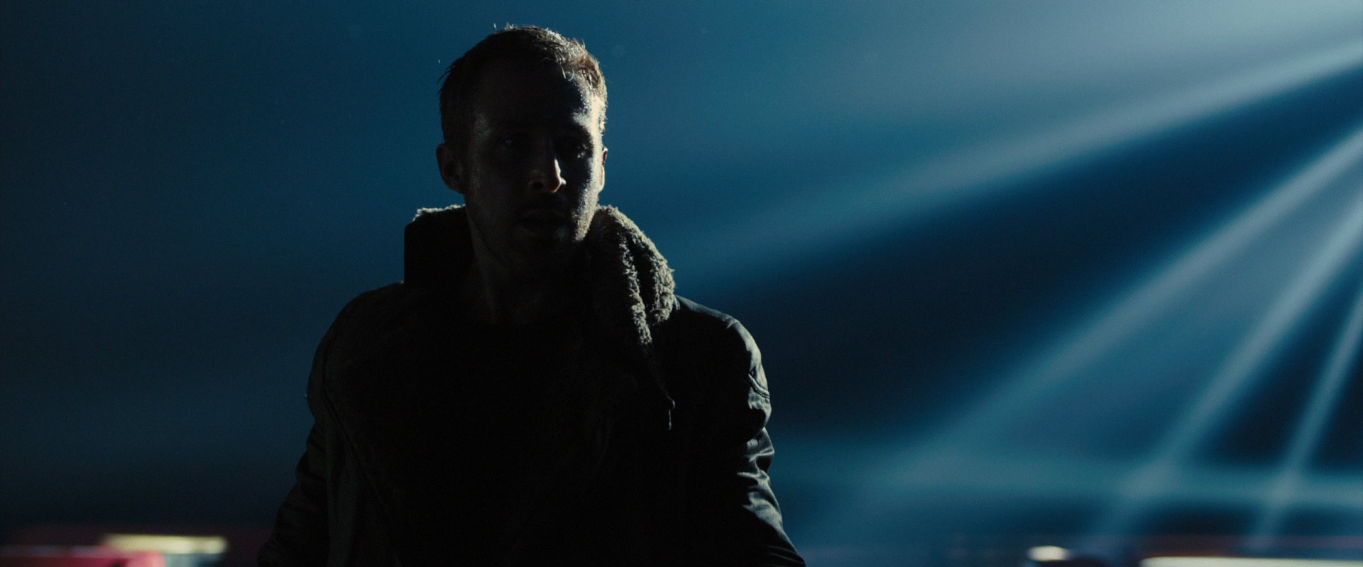 a man in a dark coat stands in a dimly lit space, with a spotlight shining on him from behind. he is backlit and his face is obscured by shadow, but his strong jawline and serious expression are still visible. the background is blurry and dark, with beams of light cutting through the darkness.
