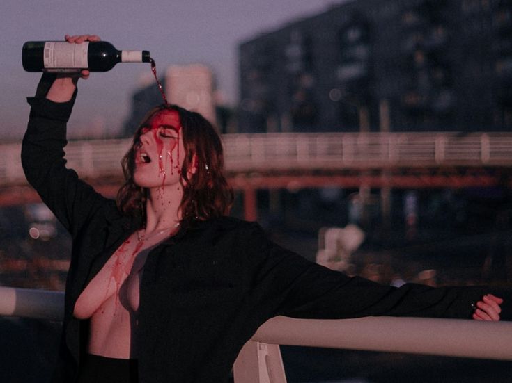 a woman stands outside in an urban setting, pouring red wine from a bottle over her head. the wine runs down her face and onto her bare chest, creating a dramatic and somewhat suggestive effect. she has her eyes closed and her mouth open, as if in ecstasy or pain. she is wearing a black jacket and is leaning against a railing. in the background, there are buildings and a bridge. the overall tone of the image is dark and artistic.