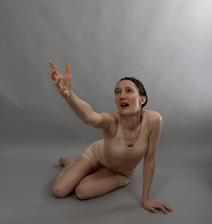 a woman is kneeling on a grey surface, looking upwards with a frightened expression. she has her hand outstretched as if reaching or pleading. she's wearing a light skincolored body suit. the background is a plain grey studio backdrop. her expression and posture convey a sense of despair and distress.