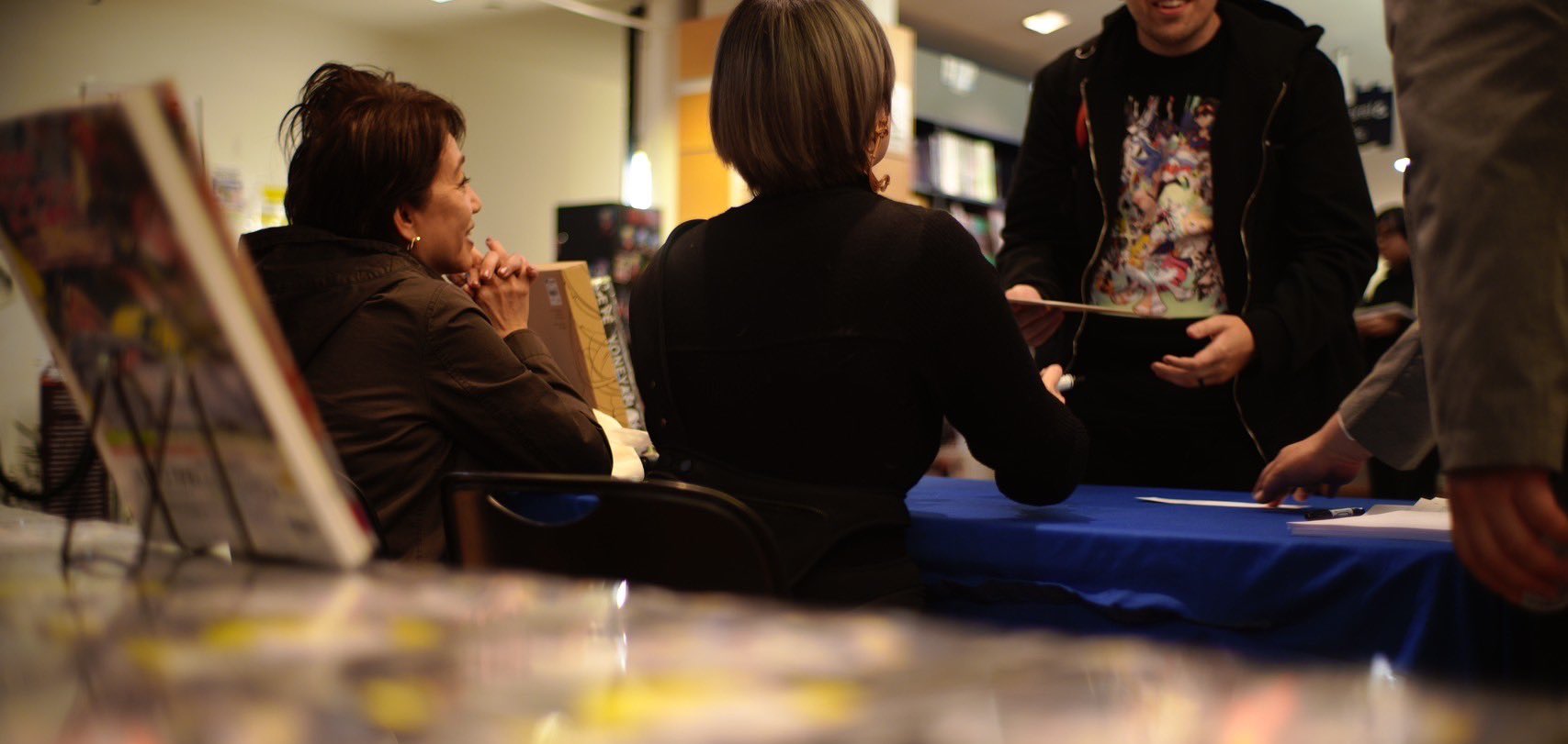 a woman sitting at a table with a man standing on the other side. the man is holding a book and appears to be talking to the woman. there are other people behind them, and it looks like they might be at a convention or book signing. the image is taken from the perspective of someone sitting at the table, looking up at the people standing.