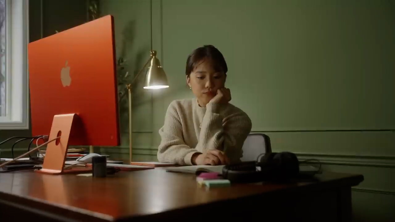 a young woman is sitting at a desk in a welllit home office. she is wearing a beige sweater and has her hand on her chin, looking intently at a notebook. on the desk is a red imac computer, a gold lamp, and a pair of headphones. the walls are painted a dark green.