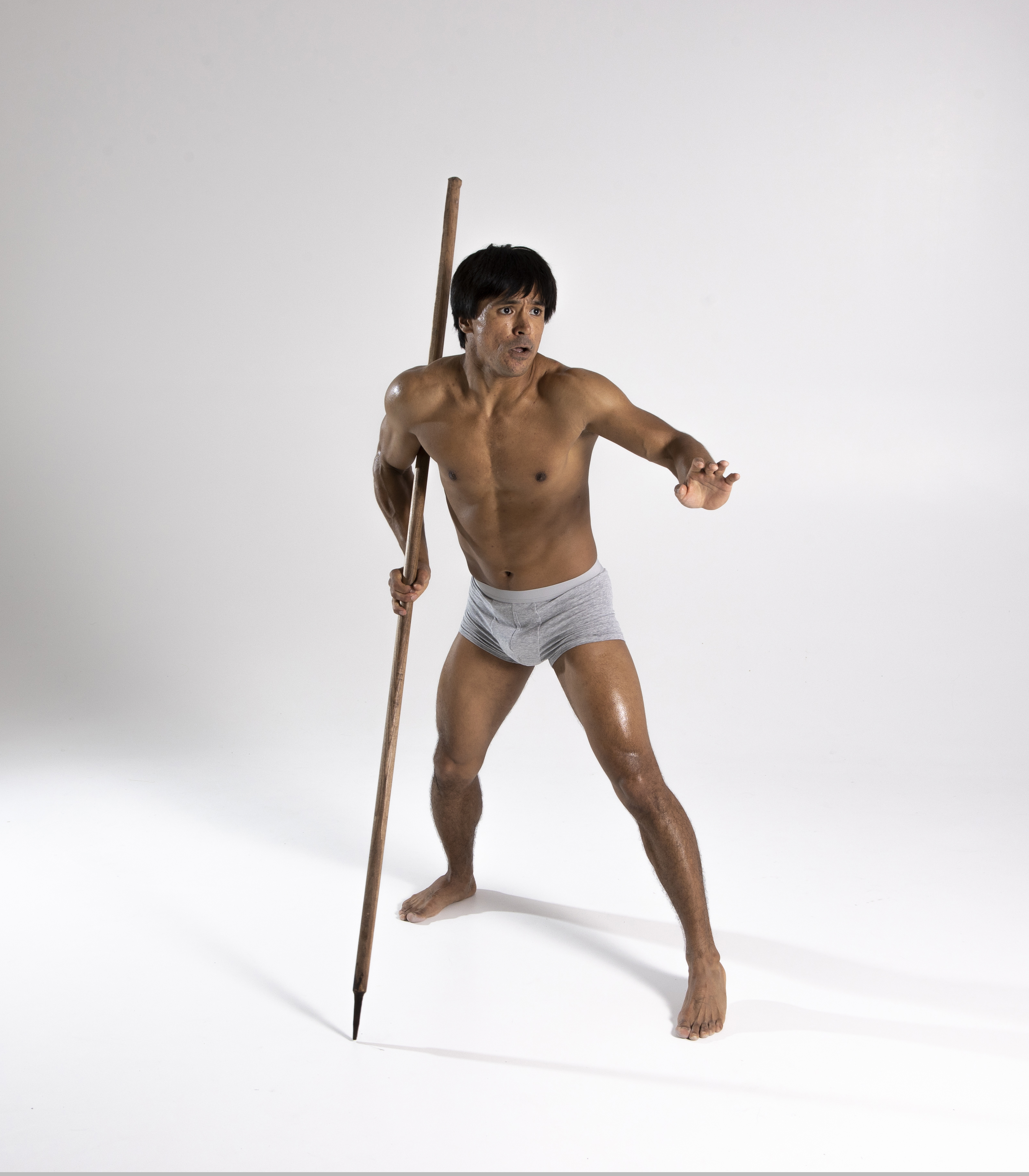 a barechested man stands in a defensive stance, holding a spear in his left hand. he wears a loincloth and looks to his right with an expression of fear or alertness. the man is wellbuilt and athletic, posing against a plain white background in a studio shot. the image captures a sense of tension and readiness for action.