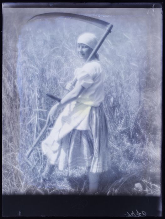 a black and white portrait of a young woman standing in a field, holding a sickle. she is wearing a white dress and a head scarf.