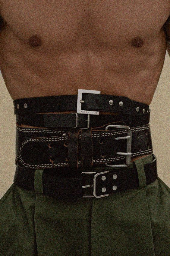 a close up view of a man's muscular torso. he is wearing a green pair of pants, and three belts around his waist. the belts are all leather and have silver buckles. the man's chest is very muscular and toned.  he is looking down at his body.