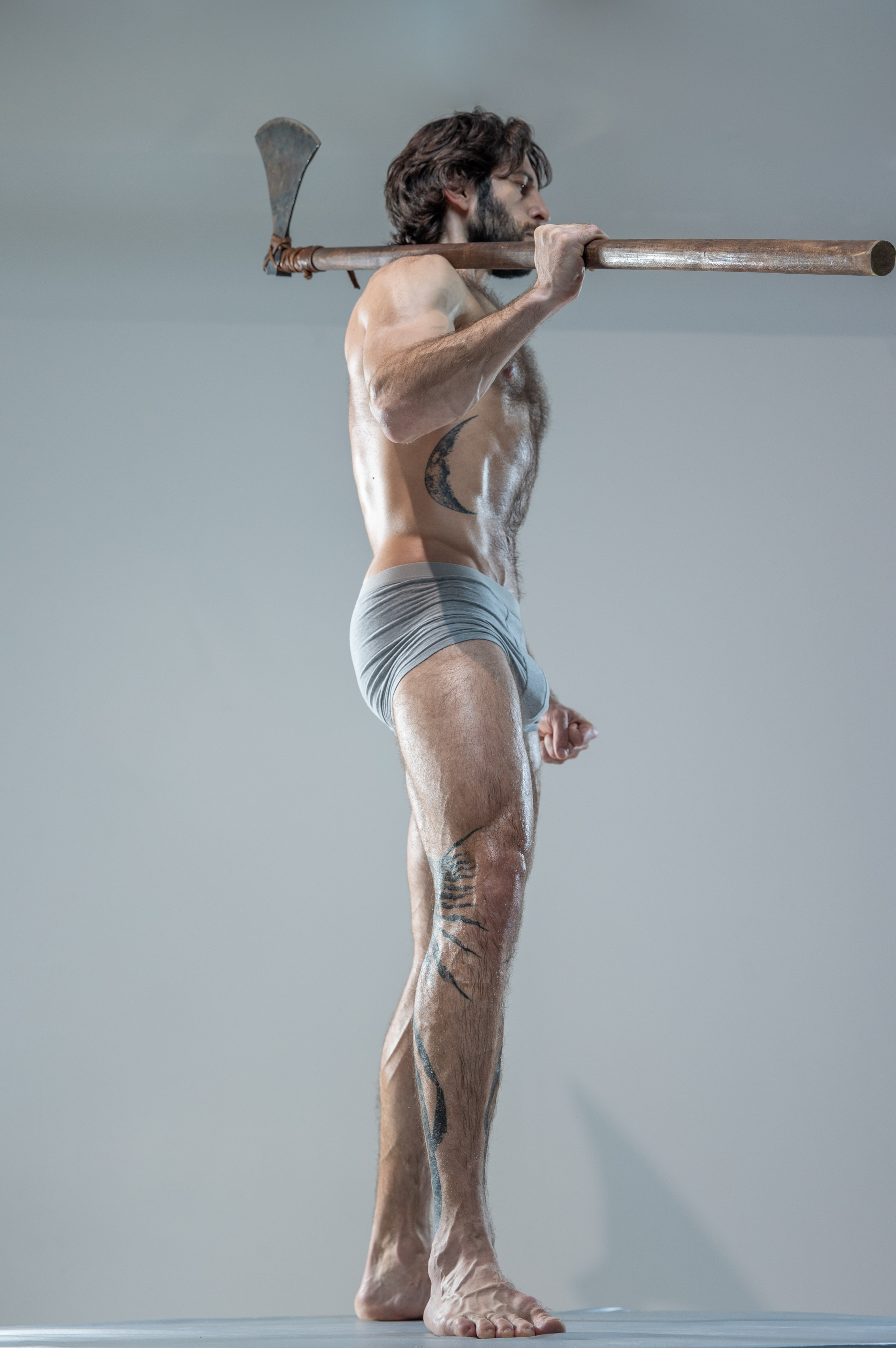 a muscular man with long hair and beard stands in a studio setting, his body slightly turned.  he holds a large, aged axe across his shoulders.  he is shirtless, wearing only gray underwear, and has several tattoos visible on his arm and leg.  his pose and expression are serious and contemplative. the image's color scheme relies heavily on grays and silvers.