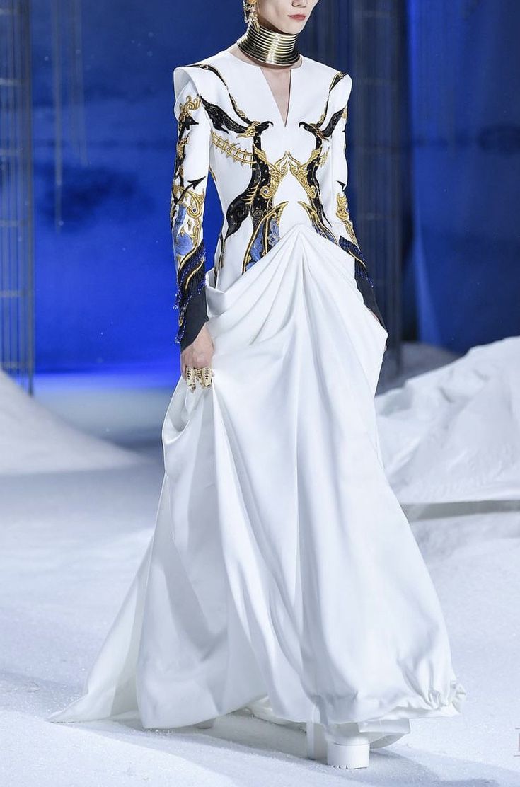 a fashion model walks the runway in a white dress with blue and gold embroidery. the dress is flowing and billowing in the wind. the model is wearing a gold collar and large gold earrings. the background is a snowy white backdrop. the overall aesthetic is wintery and elegant.