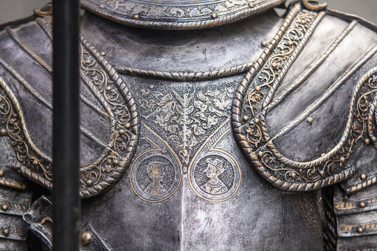 the image shows a closeup view of the torso section of a suit of medieval plate armor. the armor is made of polished metal and features intricate engravings and embossed details. two circular medallions on the breastplate depict the profiles of individuals, likely representing historical figures or the armor's original owner. the lighting is soft, highlighting the armor's metallic sheen and the depth of its engravings. the image evokes a sense of history, craftsmanship, and the weight of battle.