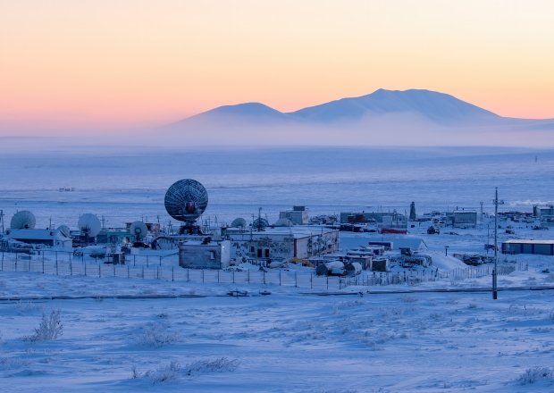 a wide shot of a snowy arctic landscape, with multiple satellite dishes and antennas. the image looks like a research or military outpost.  the sunset sky behind the facility is a deep pink color, with a mountain range visible in the distance. the snow is covering everything and it appears to be very cold.