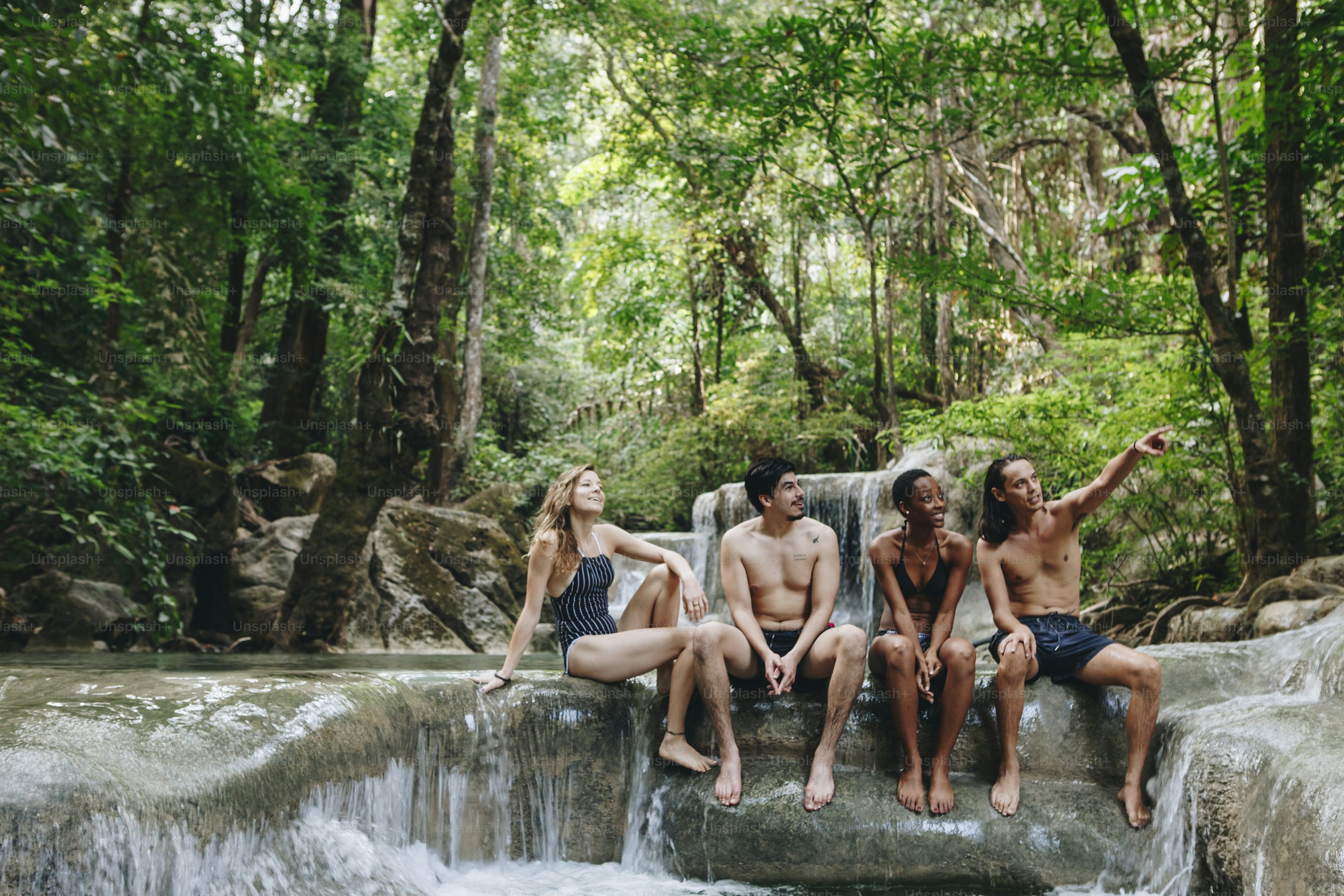 four people (two women and two men) are sitting on a rocky ledge in a lush forest, enjoying a waterfall. they are dressed in swimwear. one of the men is pointing his hand in the distance. they all appear to be happy and relaxed. the background is filled with green trees and vegetation, suggesting a tropical or exotic location.