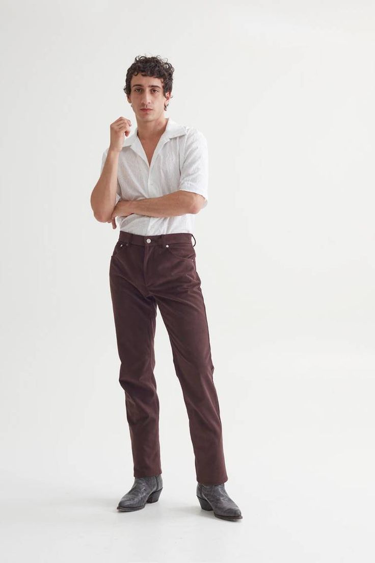 a man stands against a white background wearing brown pants, boots, and a white shirt.