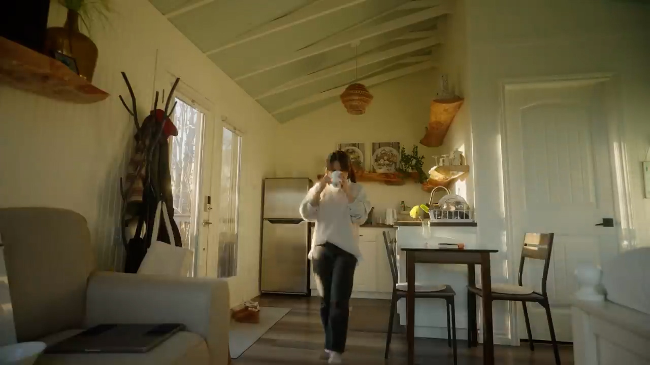 woman in a cozy, welllit tiny house enjoys a morning beverage. bathed in the warm glow of golden hour sunlight, she stands in the combined kitchen and living space. the home features minimalist decor with wooden accents and a neutral color palette.