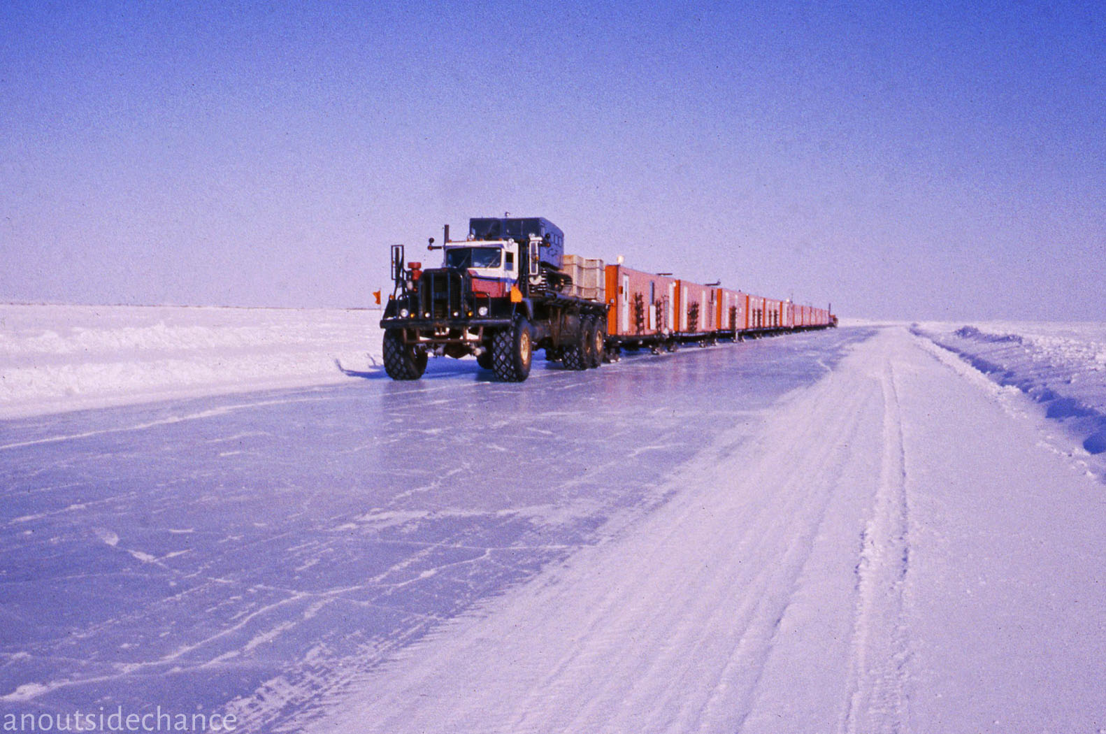 a semitrailer truck hauls a long train of flatbed trailers across a snow and ice covered road in the arctic.