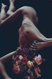 the image is an artistic nude photograph of a slender female model lying on her back. her body is arched, and she is draped with a large, lush bouquet of darkcolored flowers, mainly shades of deep red, burgundy, and pink.  the flowers partially obscure her lower body and seem to almost cradle her. the background is dark and moody, emphasizing the contrast between the model's pale skin and the rich colors of the flowers. the lighting is soft and diffused, enhancing the sensual and intimate atmosphere. the overall aesthetic is one of artistic beauty and vulnerability.
