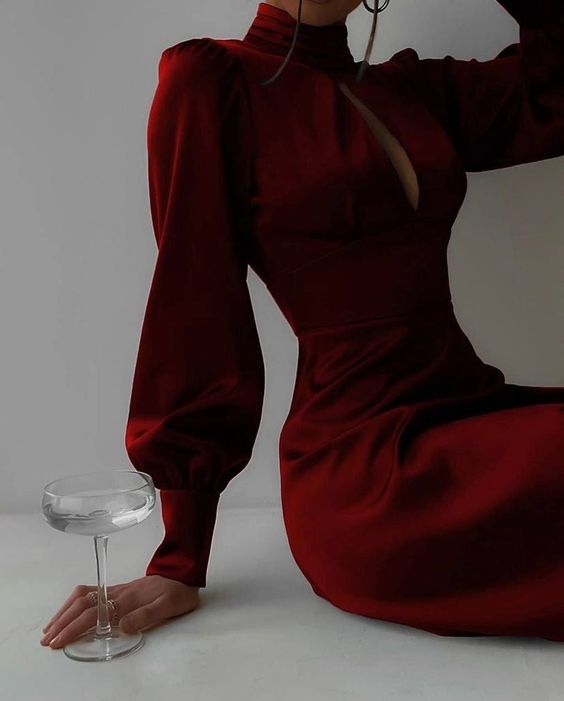 a woman is wearing a long, elegant, red silk dress with a high neck and long sleeves. she is holding a wine glass in her hand and sitting on the floor. the image is a closeup and the lighting is soft.
